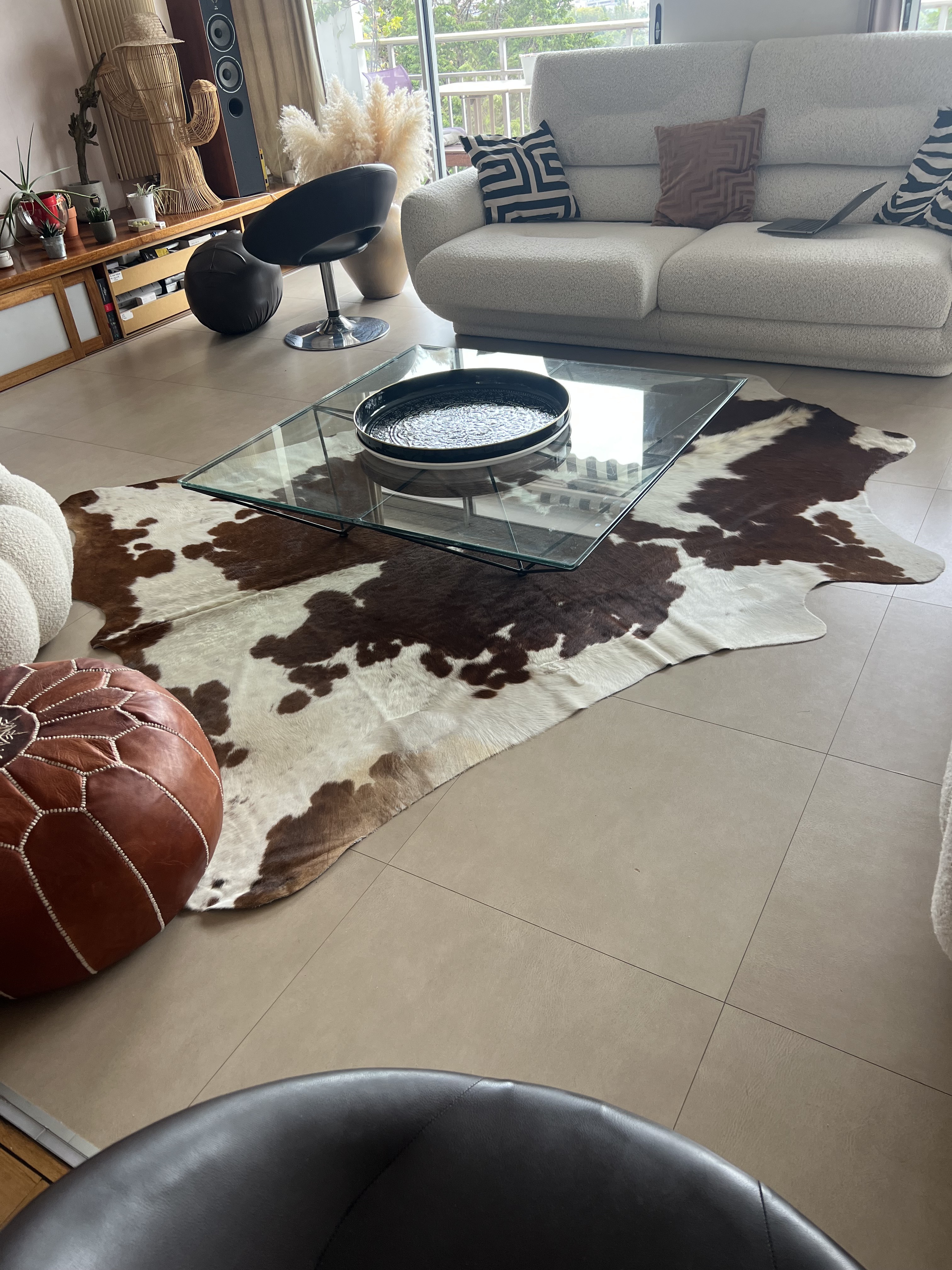 Braun-weißes Kuhfell unter einem Couchtisch aus Glas vor einem hellen Sofa auf Fliesenboden.
