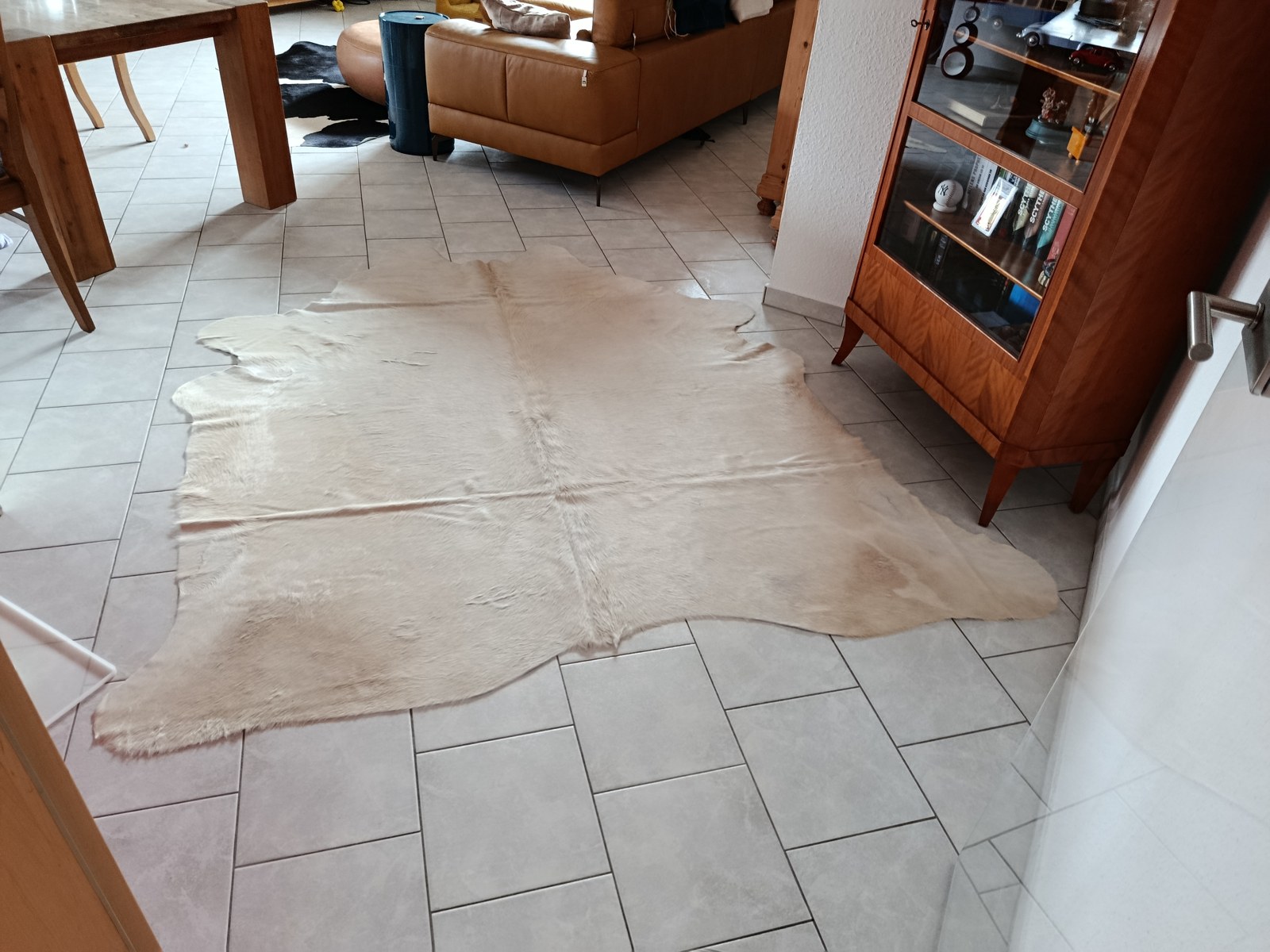 Cremefarbenes Kuhfell auf hellgrauen Fliesen in einem Wohn-Esszimmer mit Holzesstisch, cognacfarbenem Ledersofa und Holzvitrine