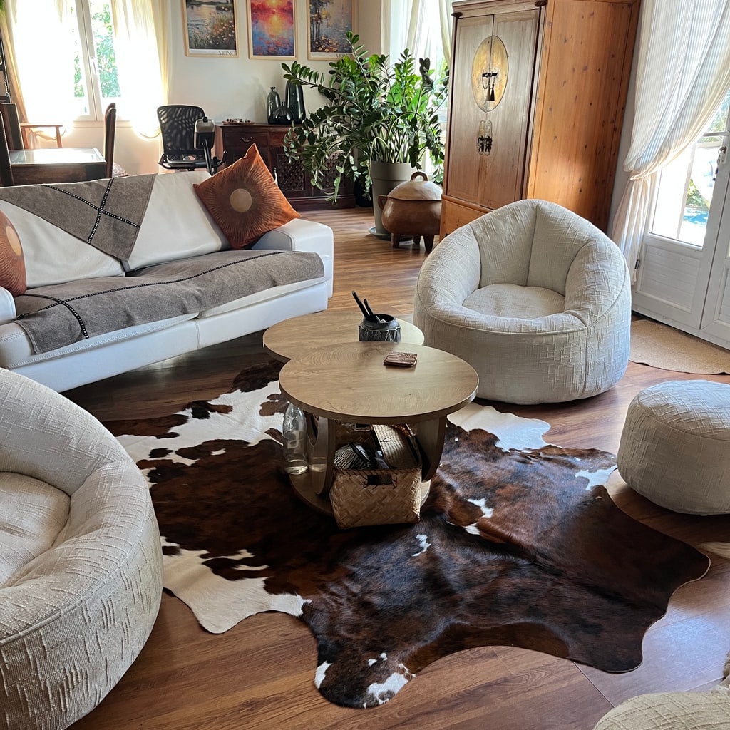 Dunkelbraun-weißes Kuhfell unter zwei Holzcouchtischen in einem hellen Wohnzimmer mit weißen Sitzmöbeln.