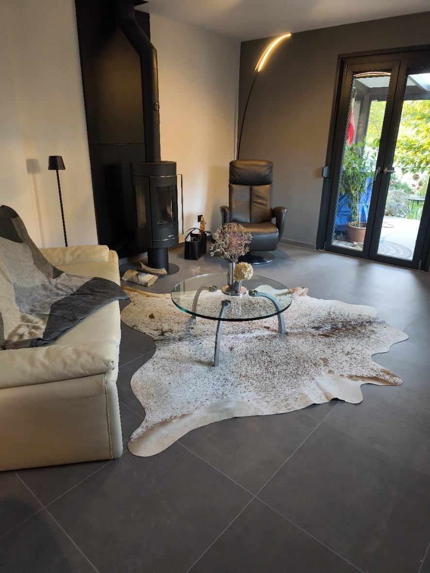 Braun-weiß gesprenkeltes Kuhfell unter einem gläsernen Couchtisch zwischen einem hellen Sofa und einem schwarzen Kaminofen.